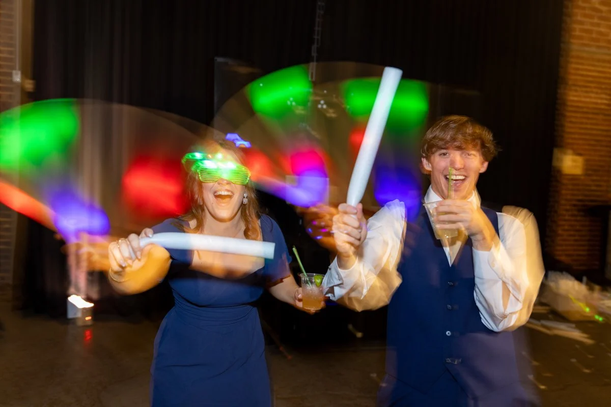 Two people celebrating with glow sticks and drinks at an indoor party, with colorful lights creating motion blur.