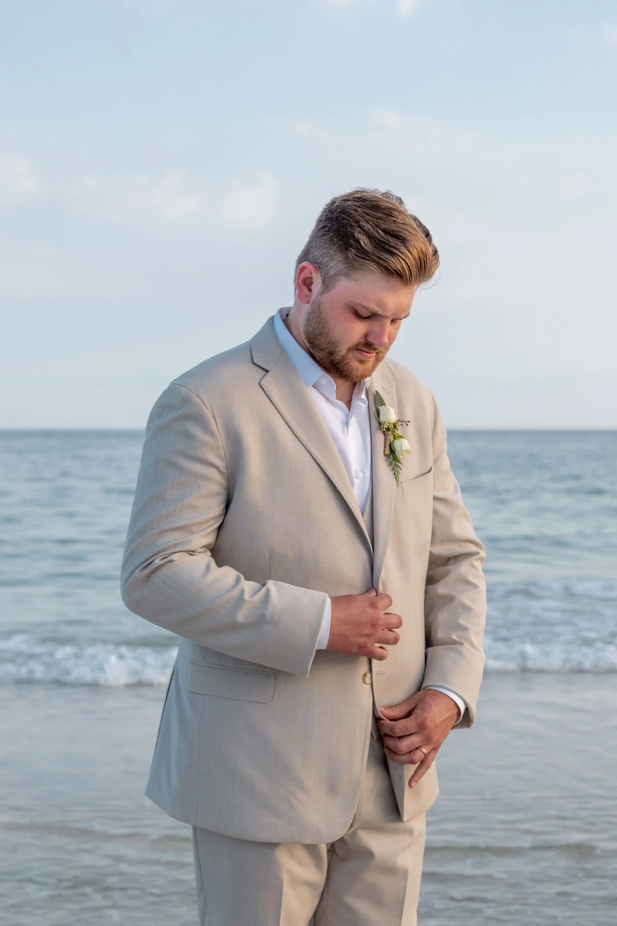 A man in a beige suit with a boutonniere stands on a beach, looking down with a contemplative expression, near the ocean under a cloudy sky.