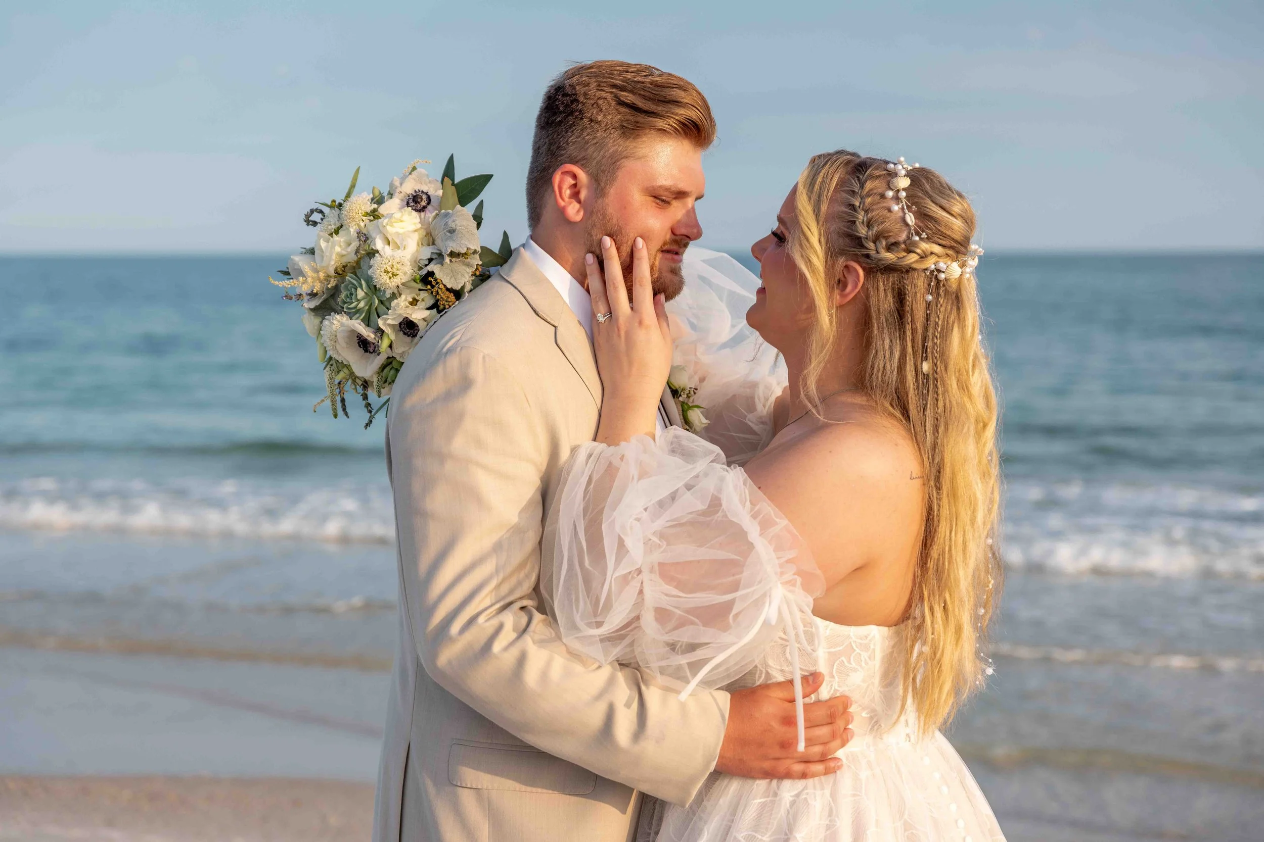 A bride and groom standing close together on the beach, embracing each other during their wedding, with the ocean and sky in the background.