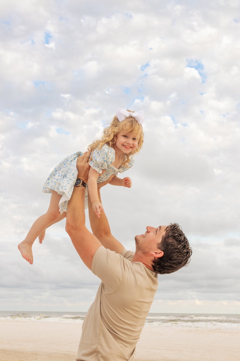 A man lifting a young girl, possibly a daughter, in the air on a beach under a cloudy sky, both smiling.