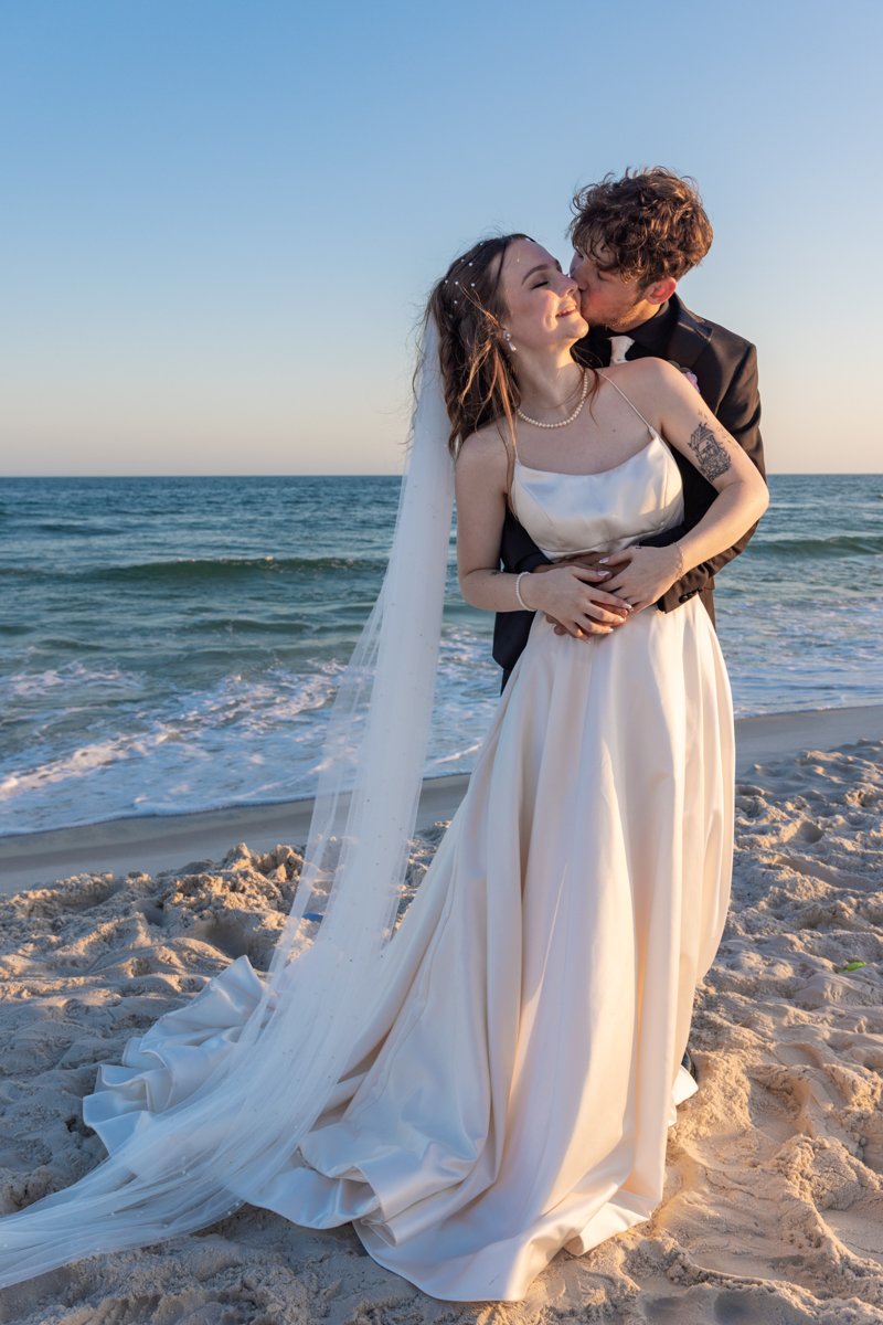 A newlywed couple sharing a kiss on the beach during sunset, with the ocean in the background, the bride wearing a white wedding dress with a long veil, and the groom in a black tuxedo.