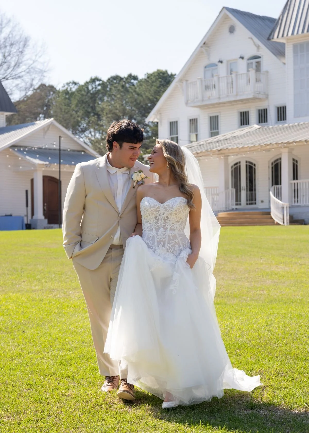 A bride and groom standing on a lush green lawn outside a white house with multiple stories and porches, smiling and looking at each other on their wedding day.