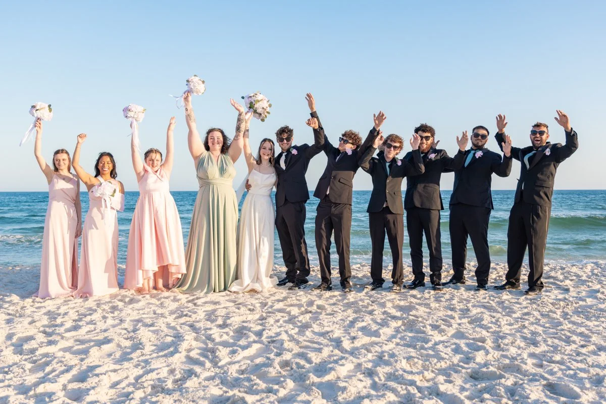 Group of wedding party members on the beach, holding bouquets, dressed in formal attire, with a blue sky and ocean in the background.