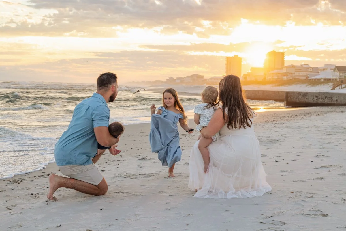 Family at the beach during sunset, with father kneeling and holding a baby, mother sitting with a toddler, and older girl playing in the sand.