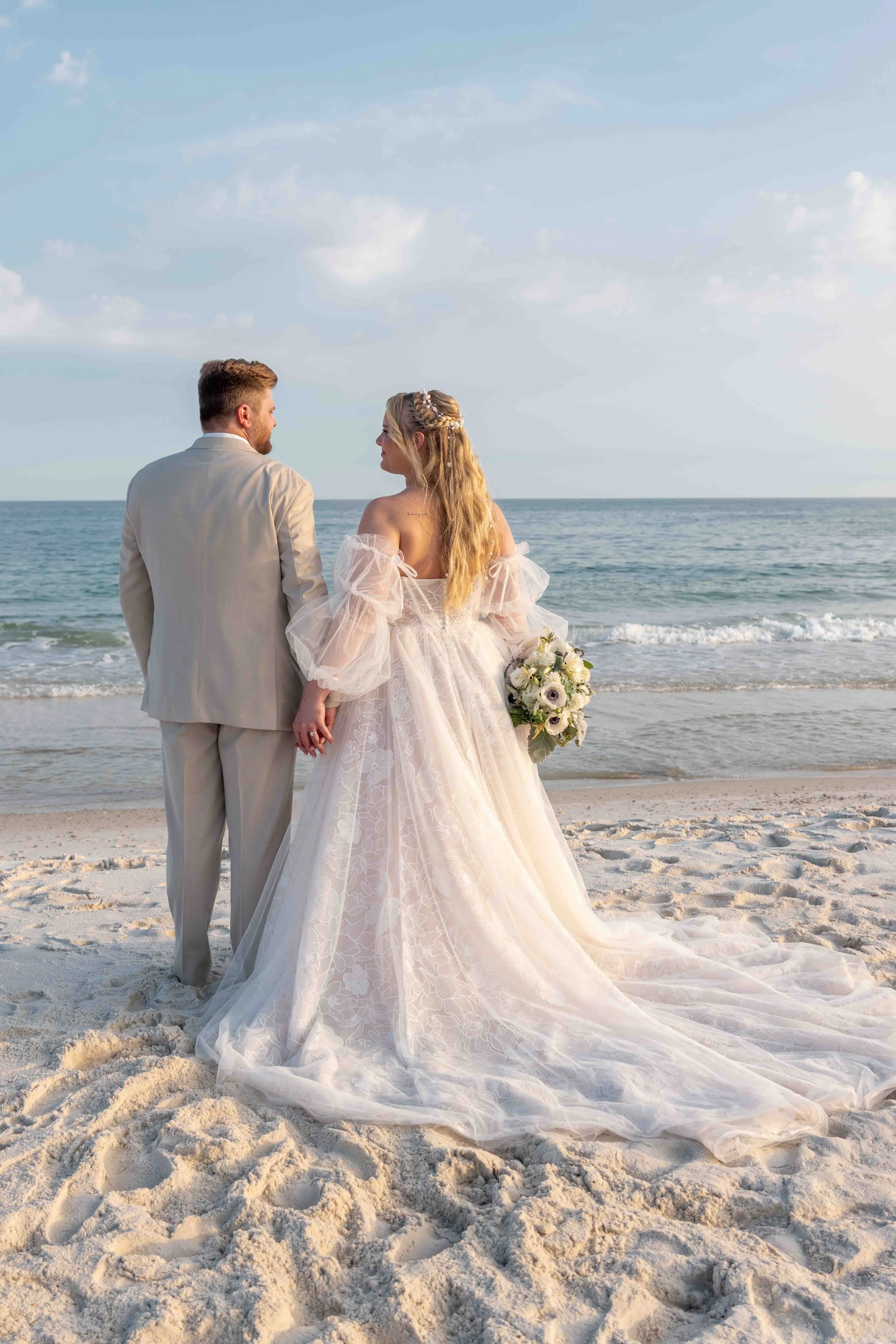 A bride and groom standing hand-in-hand on a sandy beach, facing each other, with the ocean and a partly cloudy sky in the background, during a wedding ceremony.