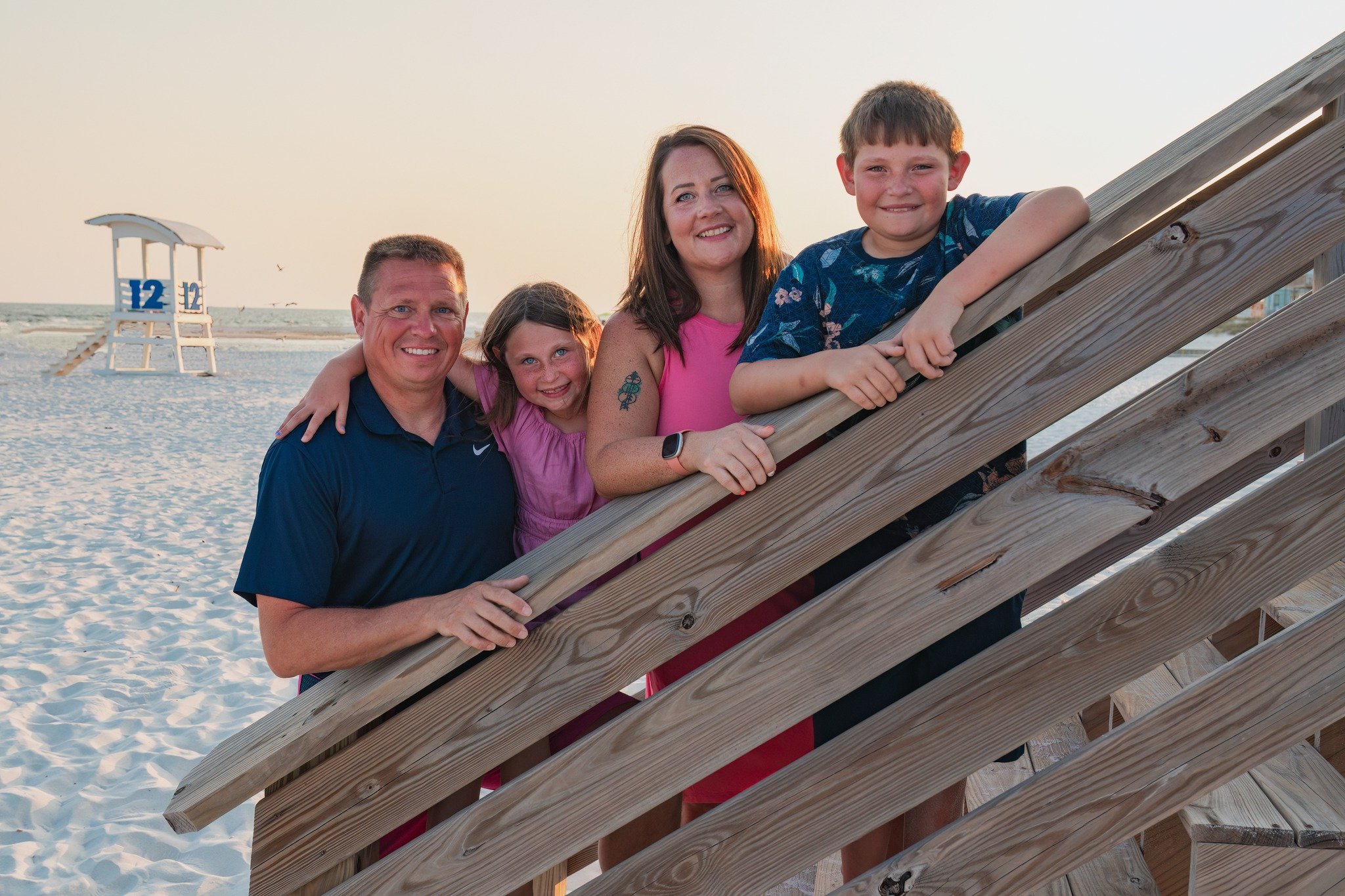 A family of five posing on a beach boardwalk during sunset, with lifeguard tower and ocean in the background.