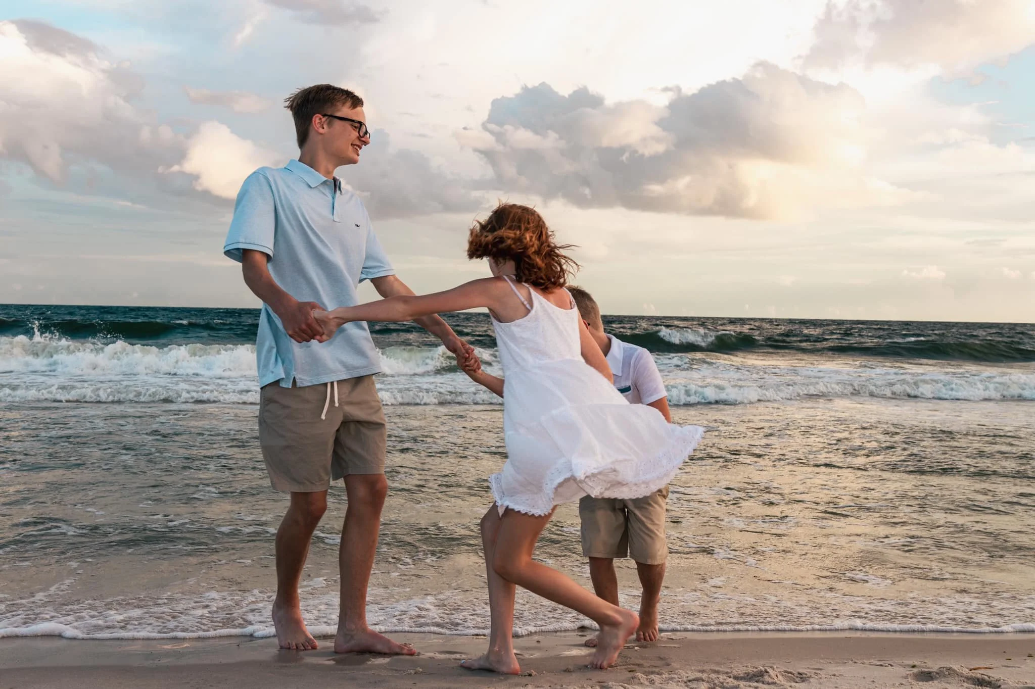 Young man and woman holding hands and playing on the beach near the ocean, with children in the background and a cloudy sky.