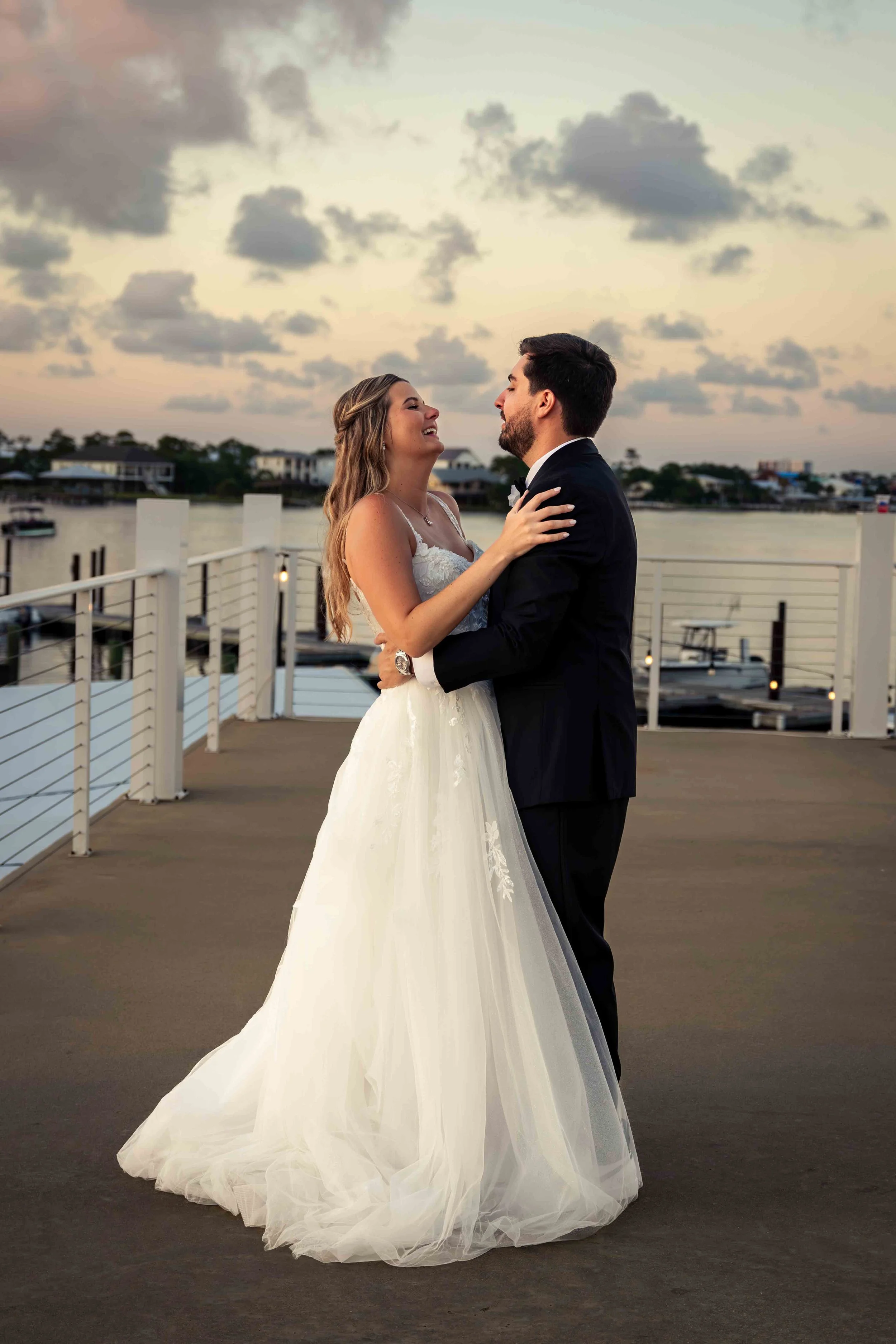 A bride and groom are dancing outdoors by the water at sunset, with the bride wearing a white wedding dress and the groom in a black tuxedo.