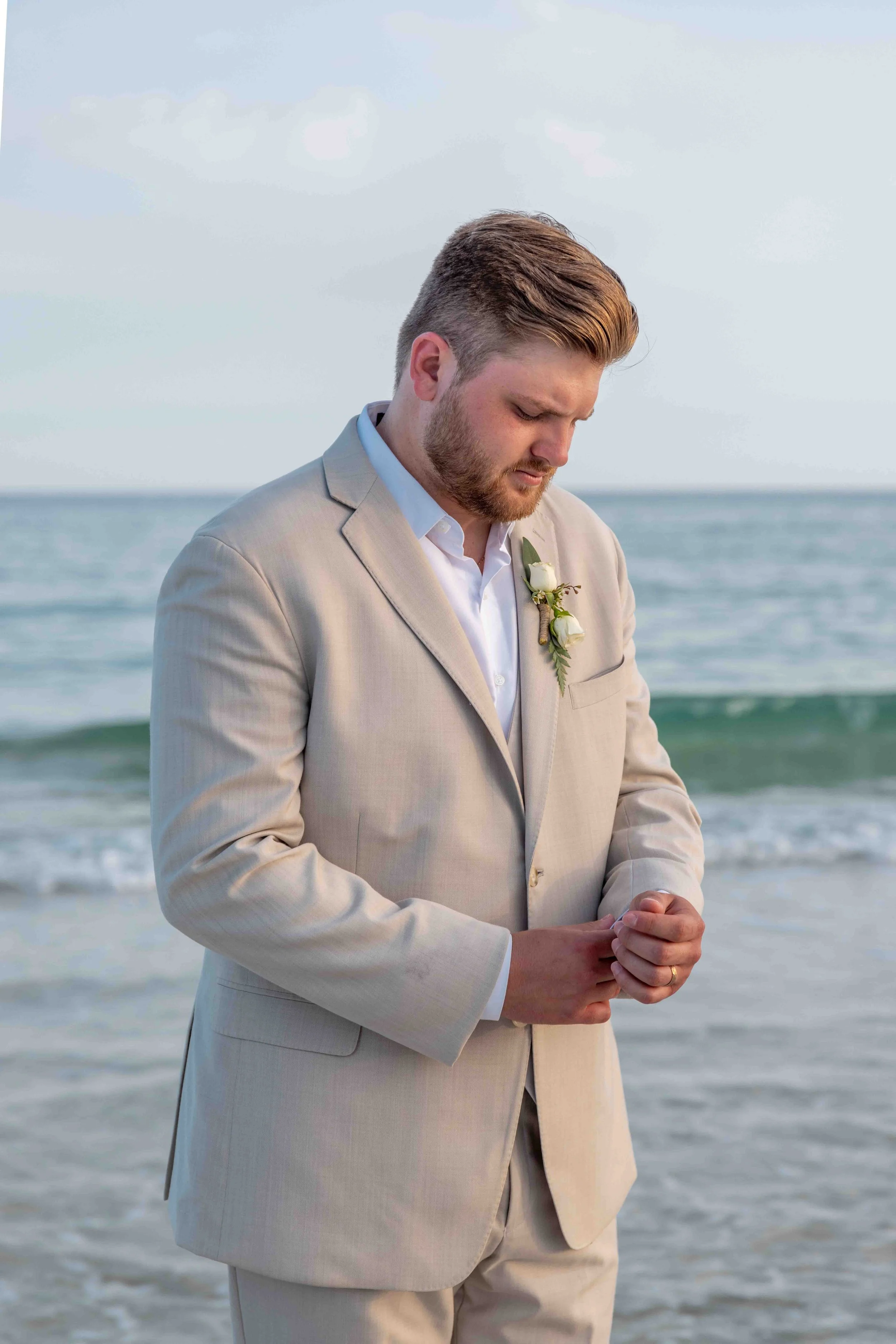 A man in a light beige suit with a white shirt and boutonnière standing on a beach, looking down with his hands clasped.