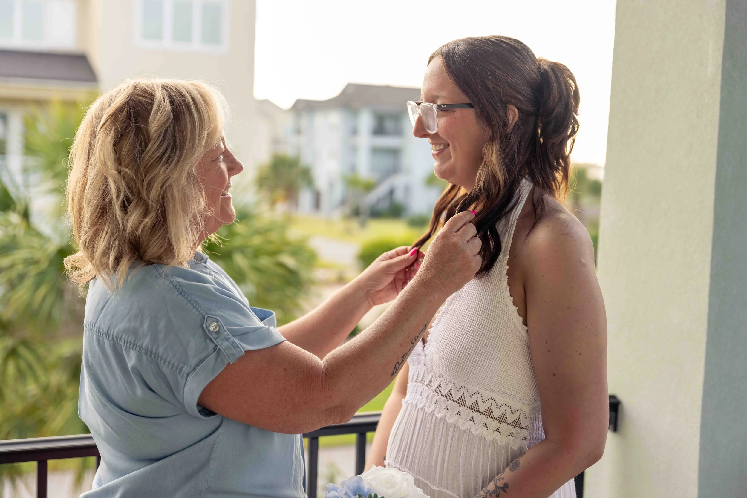 A woman with glasses and wavy brown hair smiling at another woman with blonde hair, who is wearing a light blue shirt, on a balcony with a residential neighborhood in the background.