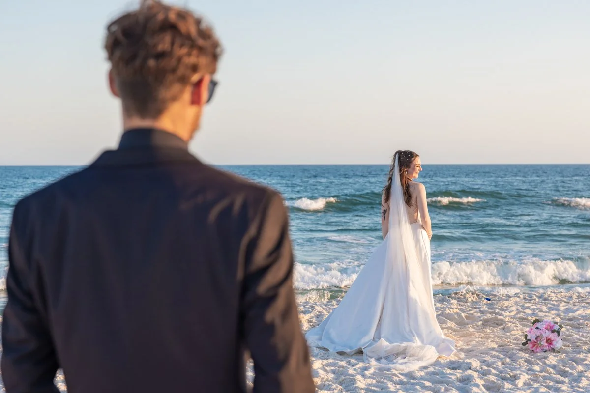 A couple in wedding attire on the beach, with the man in the foreground observing the woman standing by the shoreline, wearing a white wedding dress and veil, holding a bouquet of pink flowers on the sand.