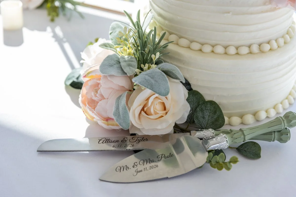 Close-up of a wedding cake with white frosting, decorative pearls, and flowers, accompanied by a cake knife and server engraved with wedding names and date, on a white table.