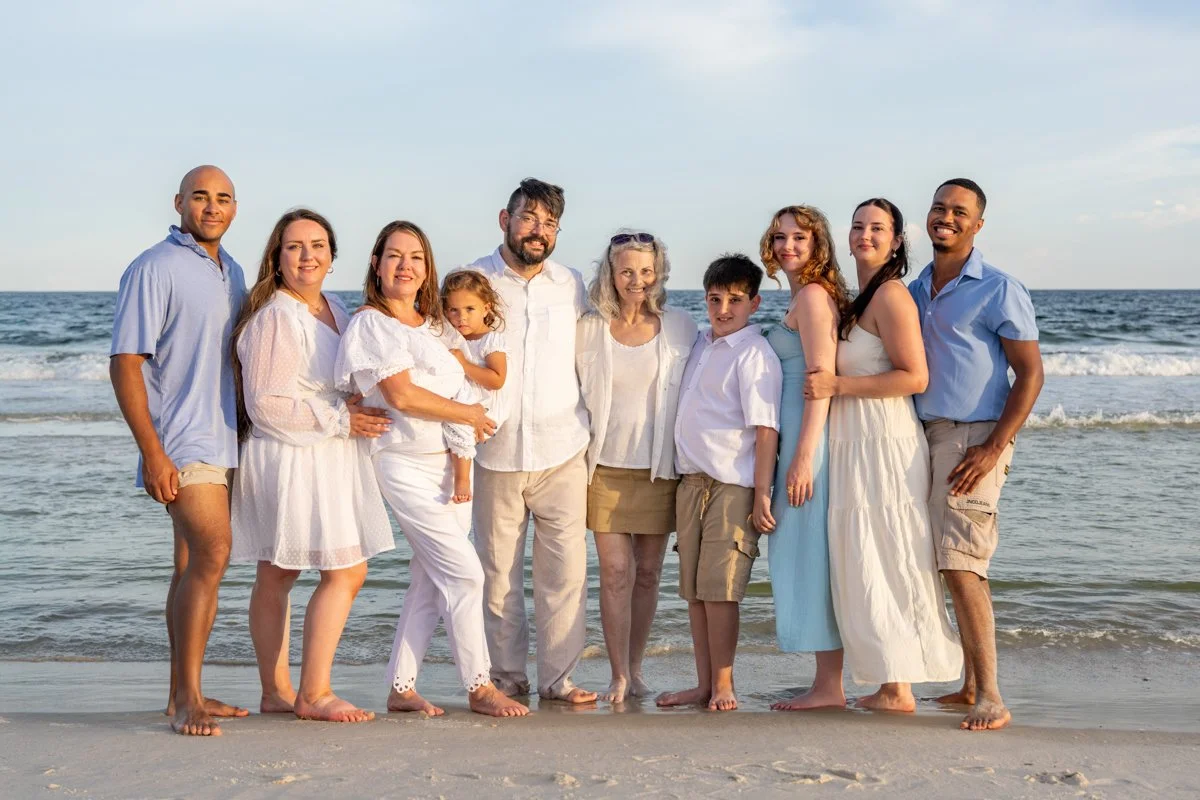 Family of ten standing on the beach by the ocean, smiling and enjoying a sunny day, dressed in light, summery clothing.