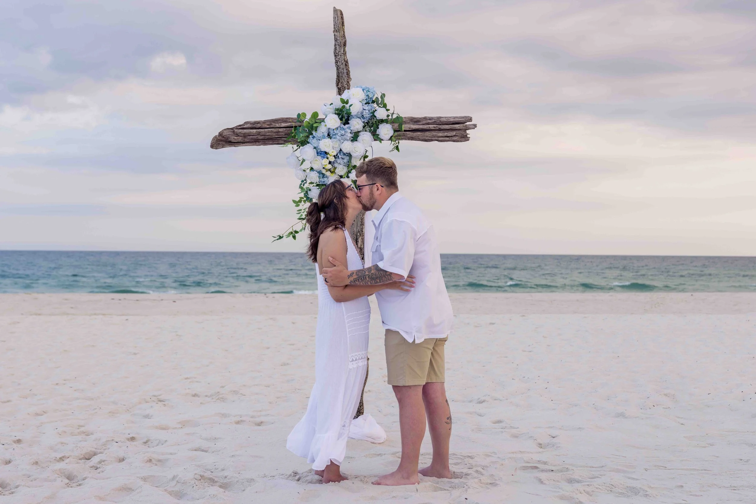 A couple in wedding attire, kissing on a beach with a large driftwood cross decorated with white and blue flowers behind them.