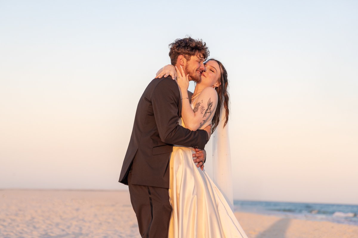 A couple in wedding attire embracing and kissing on the beach during sunset.