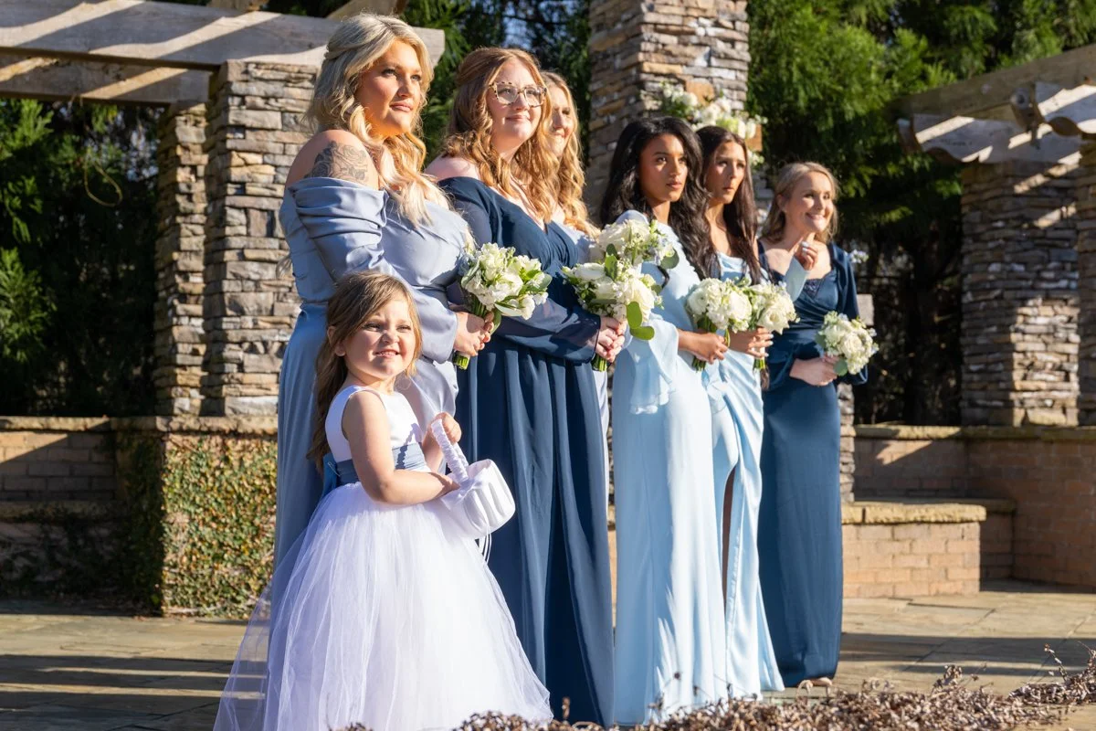 Six women and a young girl standing outdoors during a wedding ceremony, holding white bouquets, with a stone structure and greenery in the background.