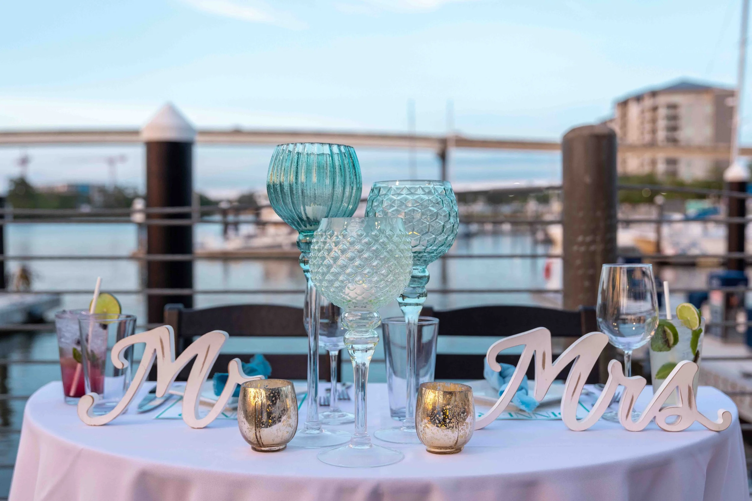 A round table with a white tablecloth decorated with several glass vases and candles, with a view of a bridge and buildings in the background, suggesting an outdoor setting for a celebration or event.
