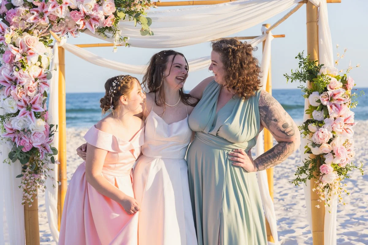 Three women standing together on a beach under a floral arch, dressed in pastel formal gowns, smiling and enjoying a moment together during a wedding or celebration on the sand with the ocean and sky in the background.