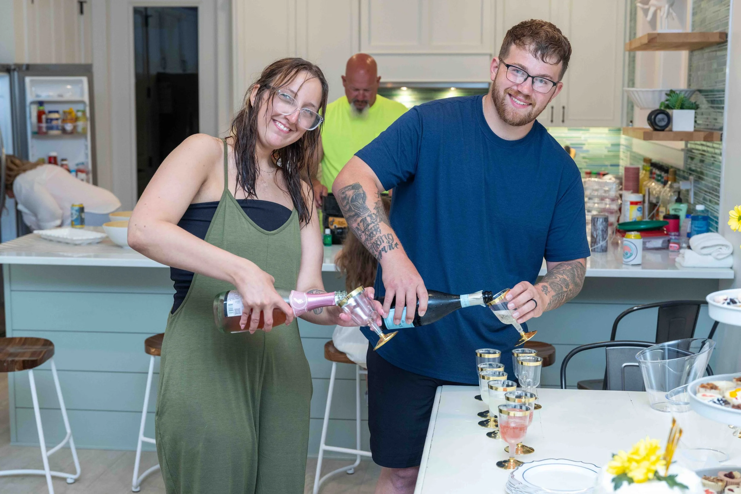Two people pouring drinks into glasses at a gathering in a modern kitchen, with a third person in the background.