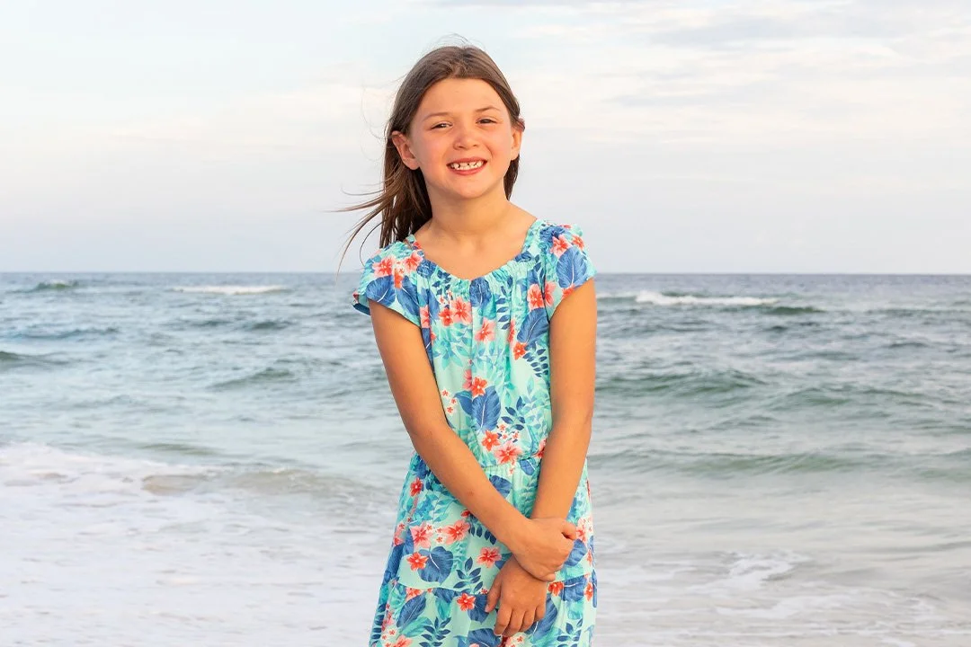 A young girl standing on the beach near the ocean, wearing a colorful floral dress and smiling.