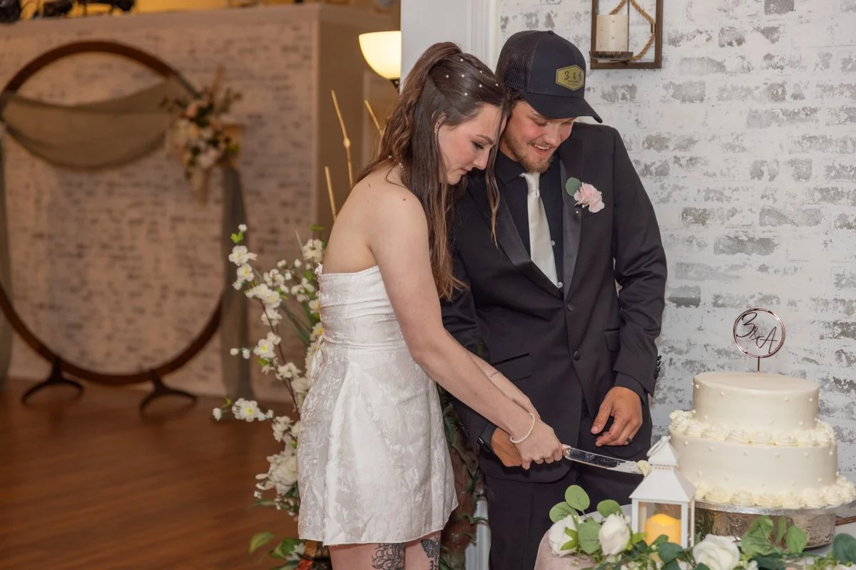 Couple cutting wedding cake, woman in white strapless dress, man in black suit and black cap, decorated wedding venue with white brick wall, floral arrangements, and a wedding cake.