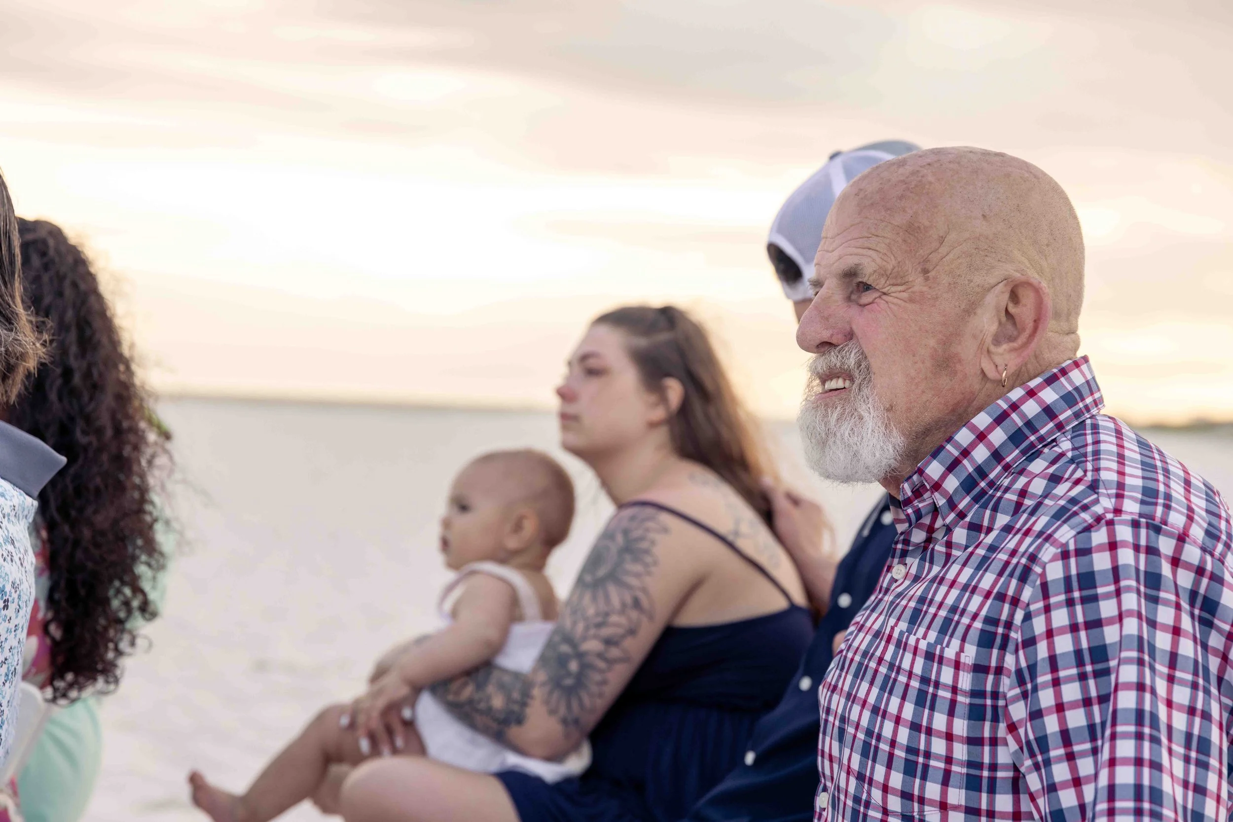 Group of people outdoors during sunset, including a smiling older man with a beard in a checkered shirt, a tattooed woman holding a baby, and two young women in the background.