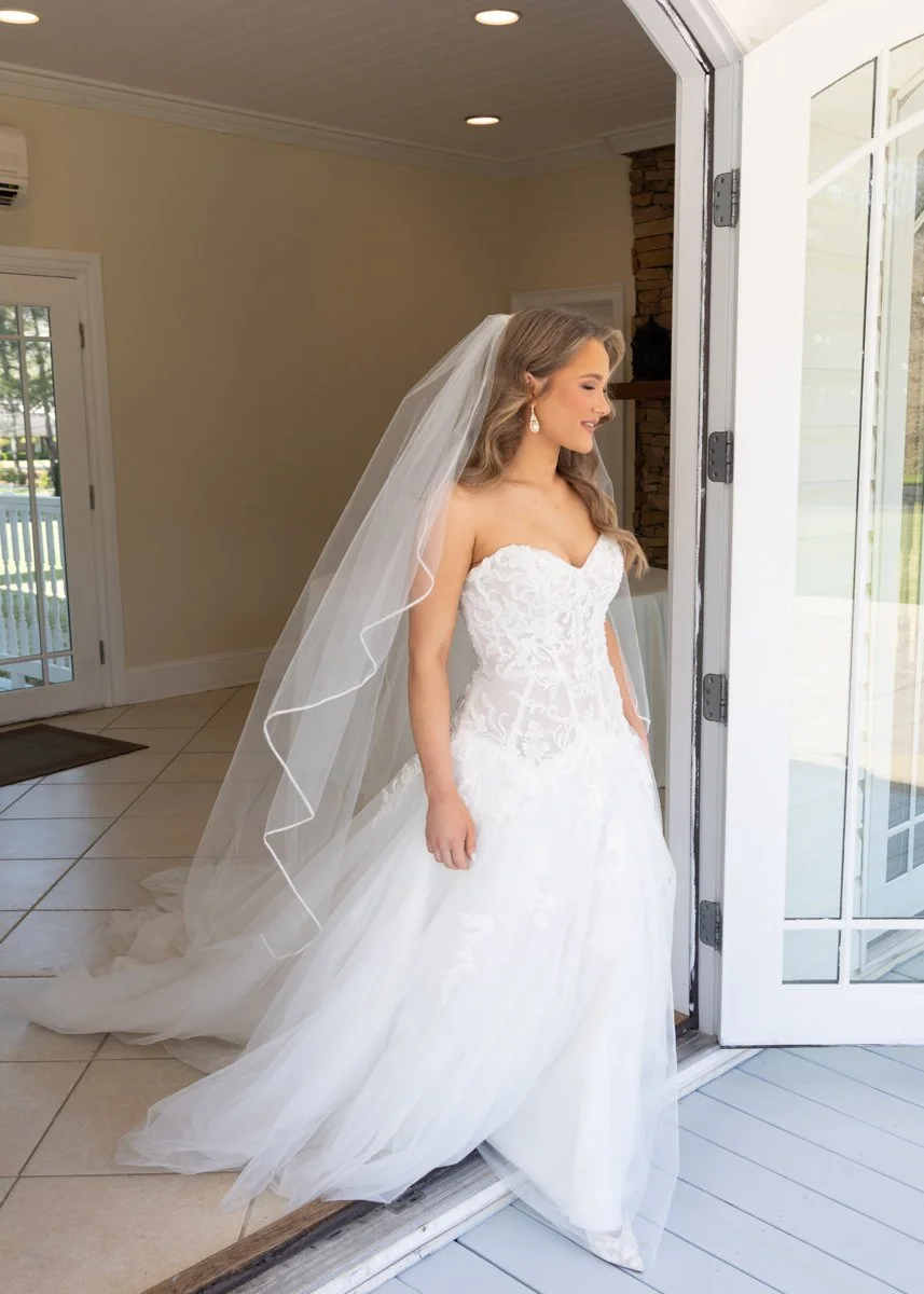 A bride in a white wedding gown with lace details, standing at a doorway, smiling and looking outside.