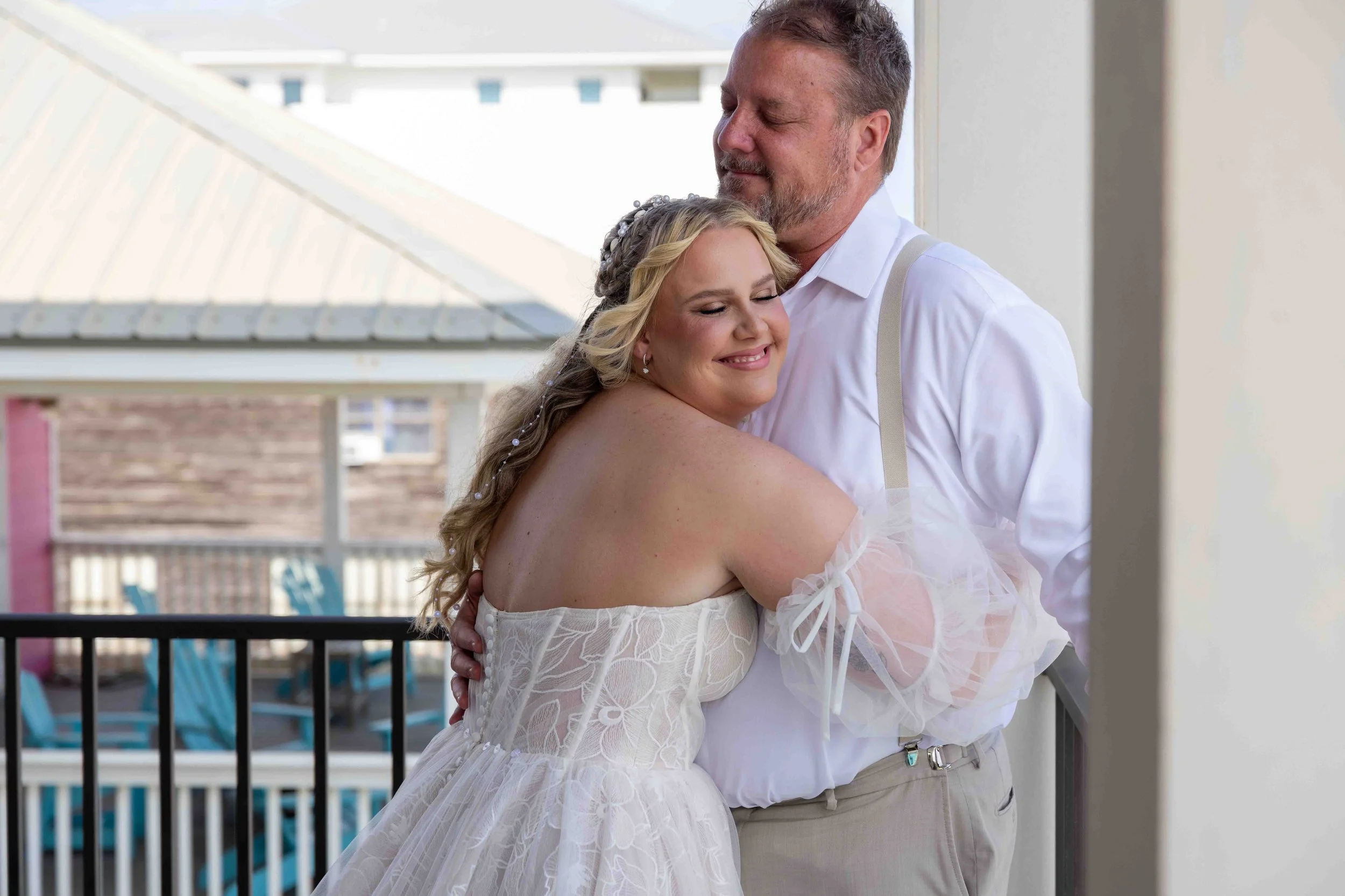 A joyful bride and groom embrace on a balcony during their wedding day, with the bride smiling and the groom gently holding her.