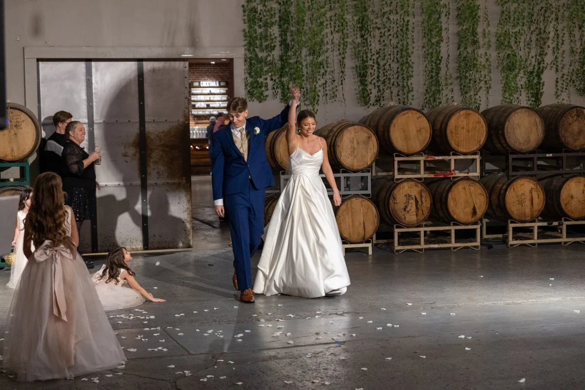 A bride and groom walking hand in hand, holding hands raised, at a wedding reception in a rustic venue with barrels and greenery in the background. Guests are watching and celebrating.