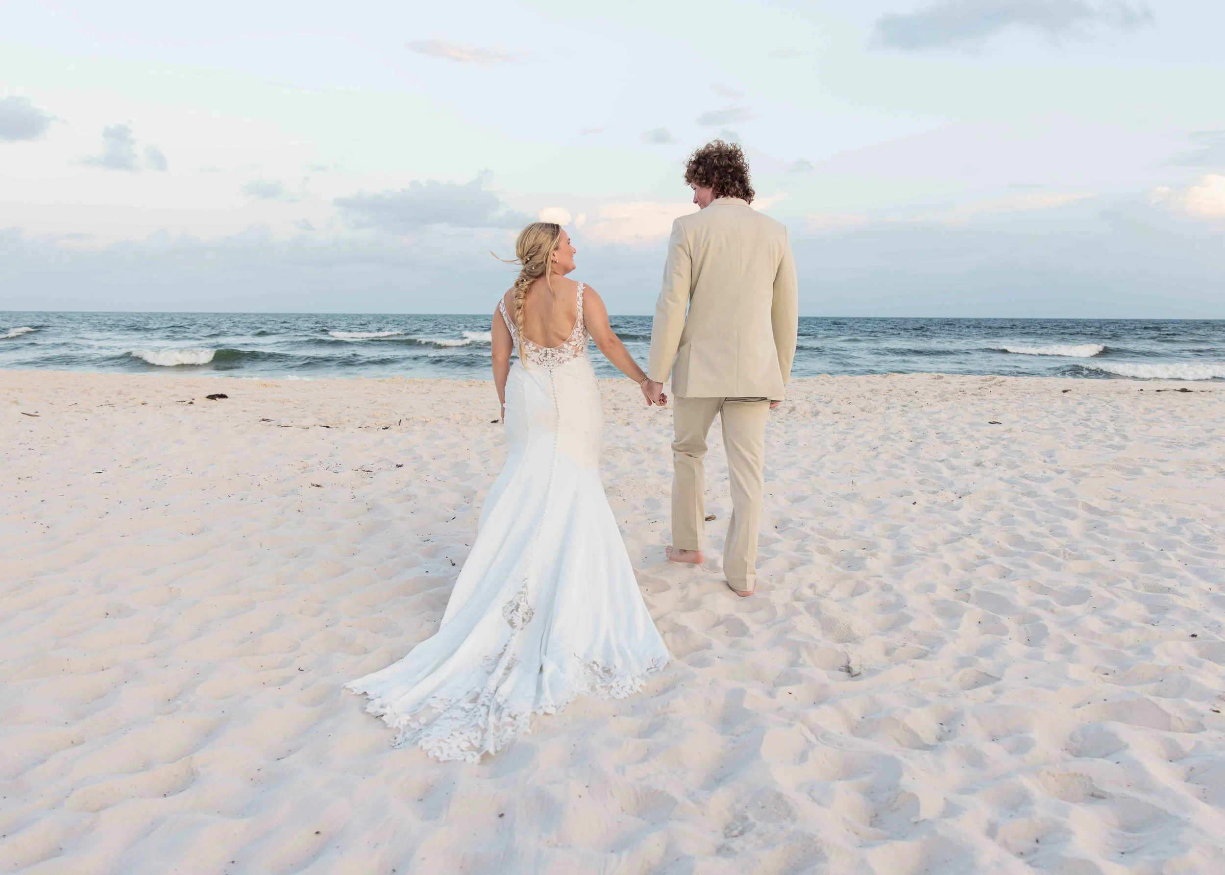 A bride and groom holding hands walking on the beach during their wedding, with the ocean and sky in the background.