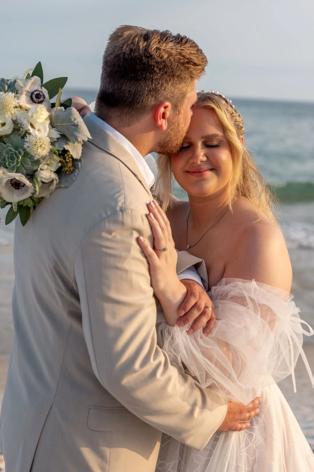 A newlywed couple embracing on the beach, with the ocean in the background. The man is kissing the woman's forehead, and she has a content smile. The man is wearing a beige suit, and the woman is in a white dress holding a bouquet of flowers.