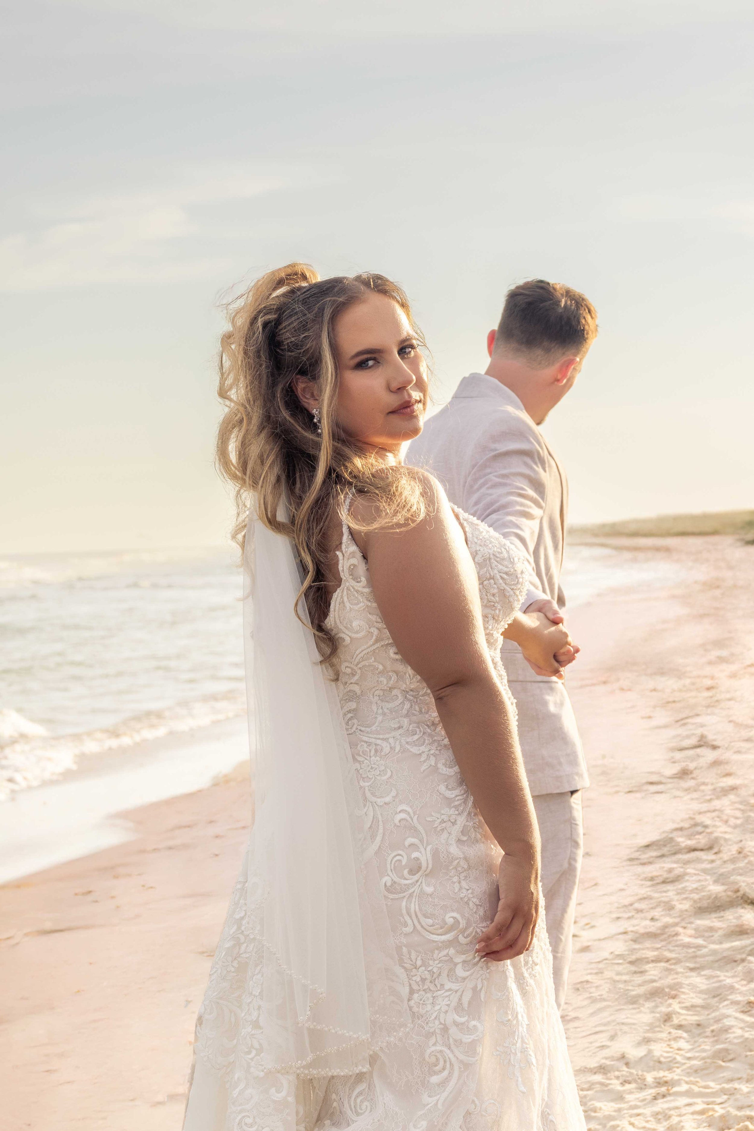 A bride in a lace wedding dress standing on the beach, looking at the camera, with a groom in a light-colored suit facing away in the background during sunset.