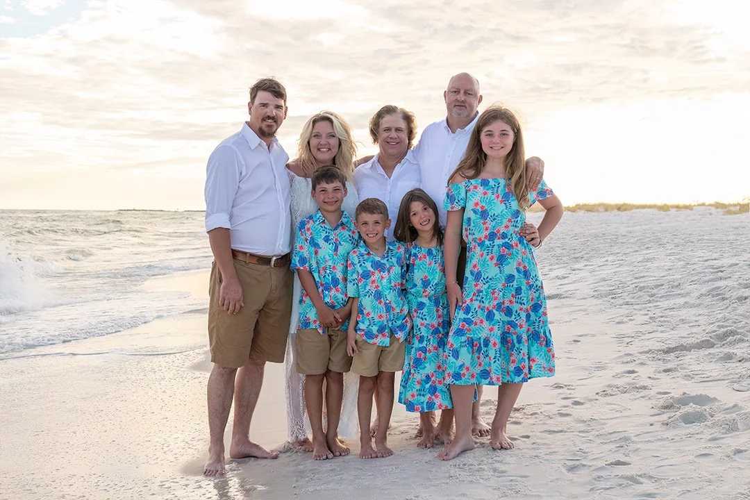 A family of nine standing on a beach at sunset, with four adults and five children, all smiling and dressed in summer clothing.