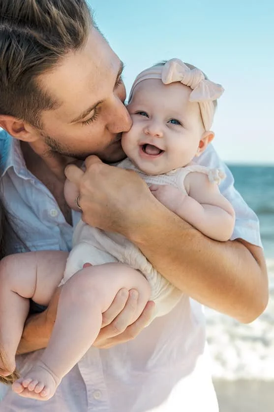 A man holding and kissing a smiling baby girl with a bow headband on a beach.