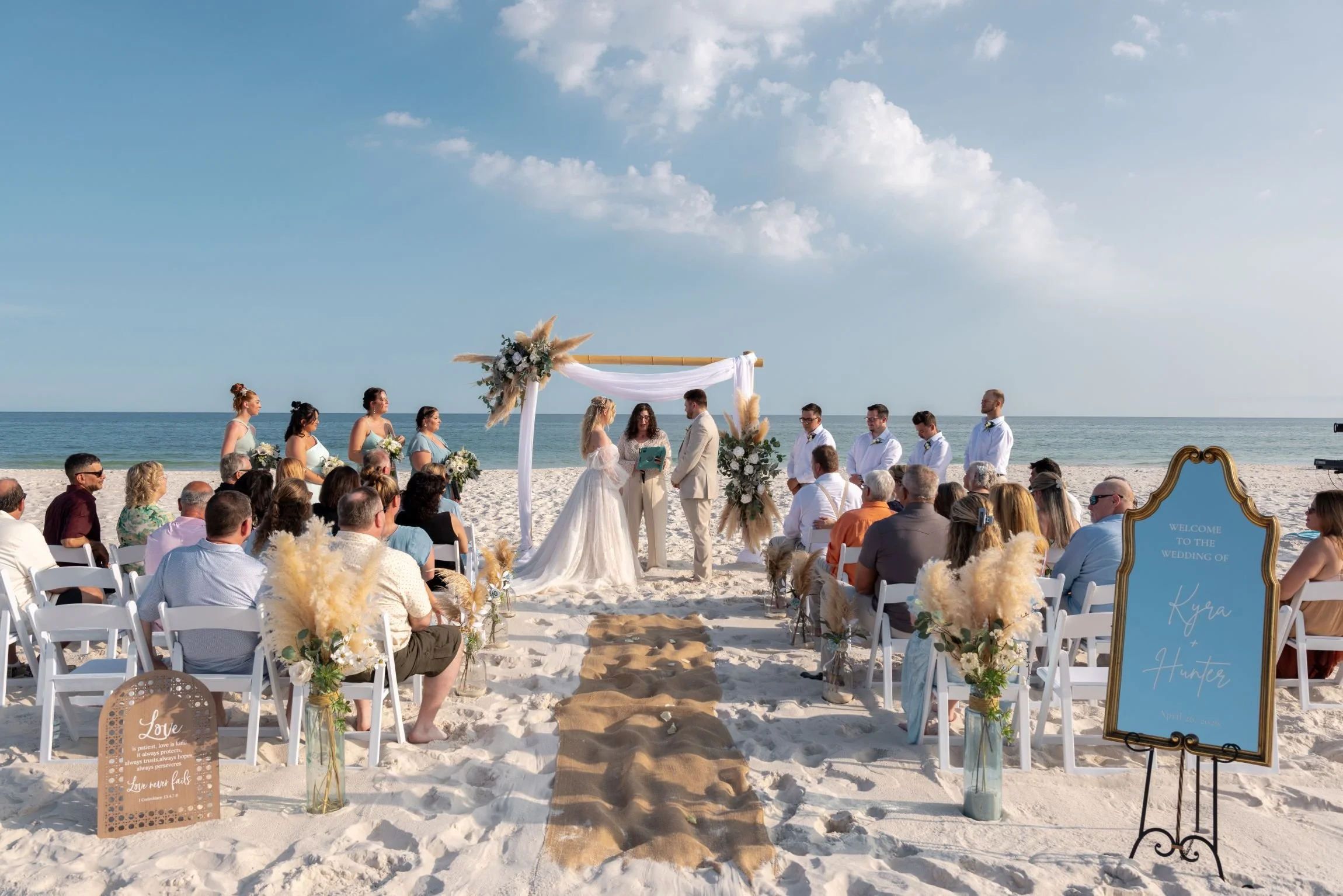 A beachside wedding ceremony with a bride and groom exchanging vows under a decorated arch, surrounded by guests seated in white chairs and floral arrangements, with the ocean and sky in the background.