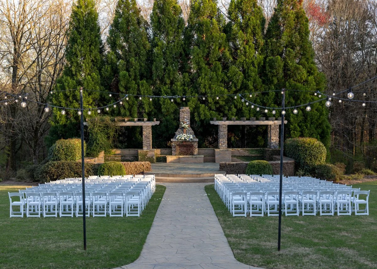 An outdoor wedding ceremony setup with white chairs facing a brick fireplace on a stage, flanked by pergolas and decorated with string lights, surrounded by lush green trees.