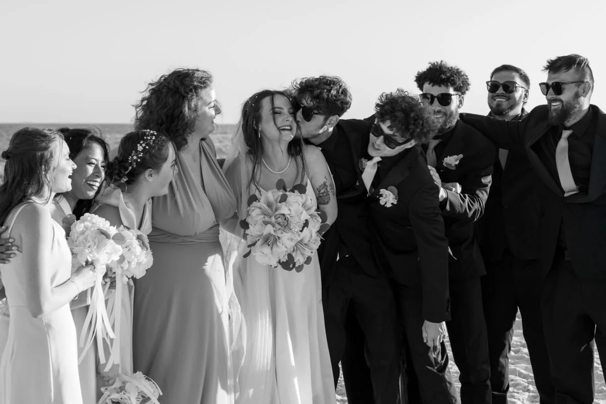 A group of nine people, including a bride and groom, celebrating on the beach. The bride is holding a bouquet, and everyone is smiling and laughing. The scene is in black and white.