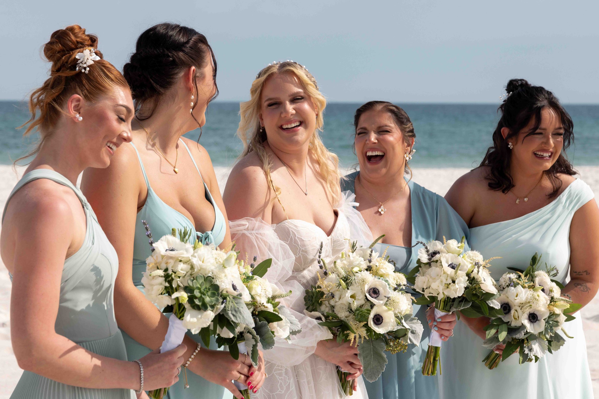 Group of women in dresses holding bouquets, standing on a beach with the ocean in the background, smiling and laughing.