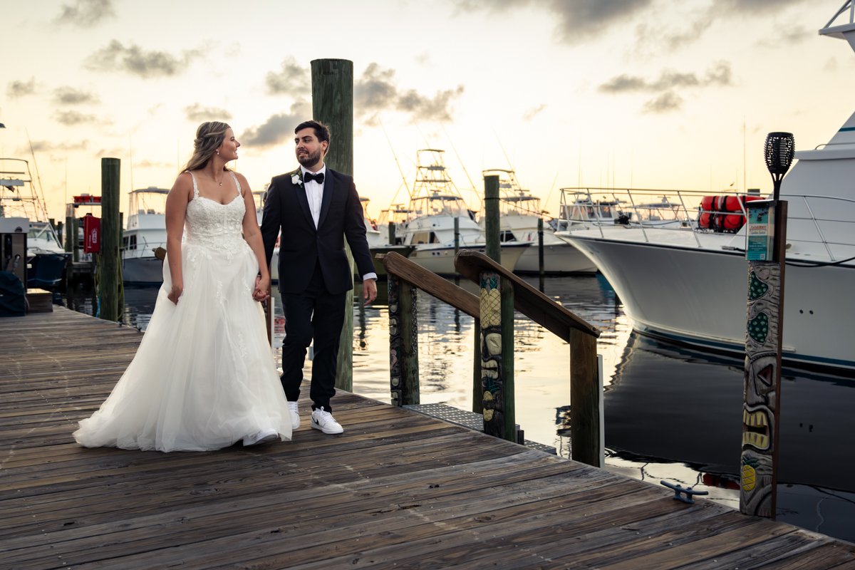 A bride and groom walking hand in hand on a wooden dock by the water with boats anchored in the background during sunset.