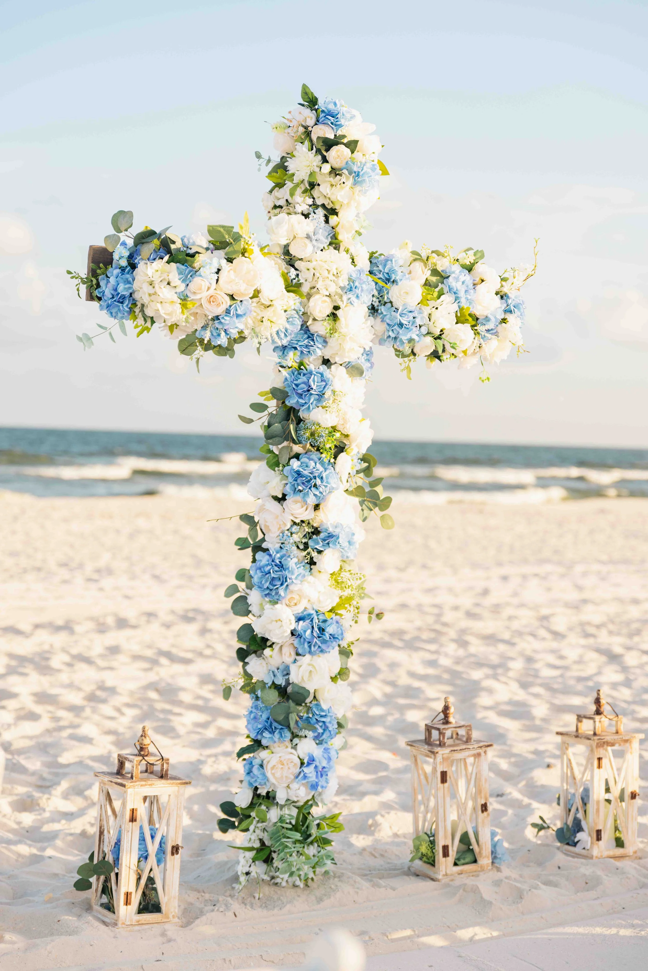 A wedding altar decorated with a large floral cross made of white and blue flowers and greenery, set up on a sandy beach with lanterns at the base, and an ocean view in the background.