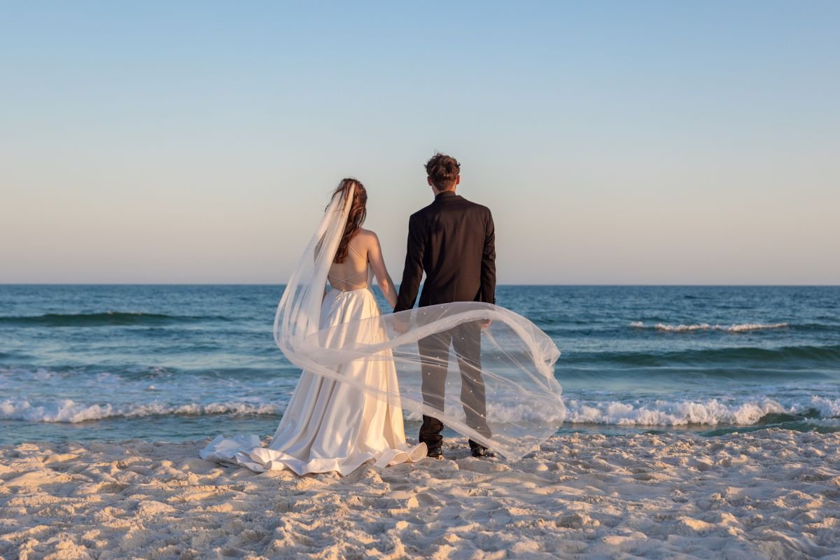 Bride and groom standing on a sandy beach facing the ocean, holding hands, with the bride wearing a long white wedding dress and veil, and the groom in a dark suit, during sunset or sunrise.