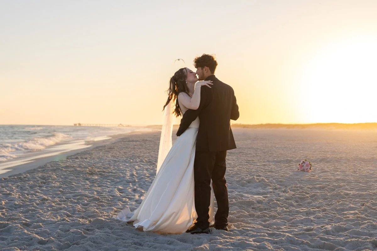 A couple in wedding attire, embracing on the beach at sunset, with a small bouquet of flowers on the sand nearby.