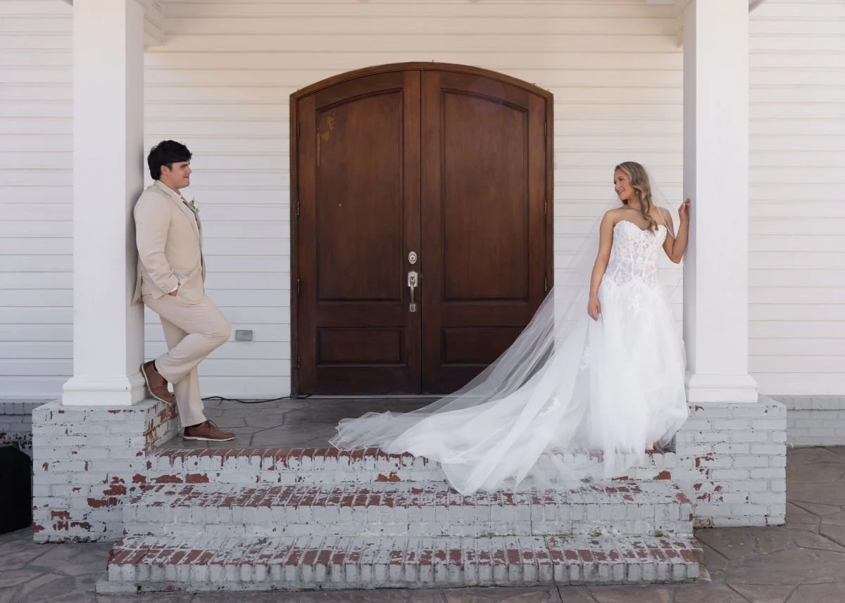 A bride and groom on the porch of a house, facing each other. The groom leans casually against a white pillar with his hands in his pockets, smiling at the bride, who stands in her wedding gown with a long train, leaning against the house wall and lo