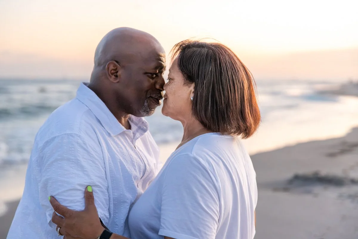 An older couple stands close together on a beach at sunset, touching foreheads and smiling, wearing white shirts.