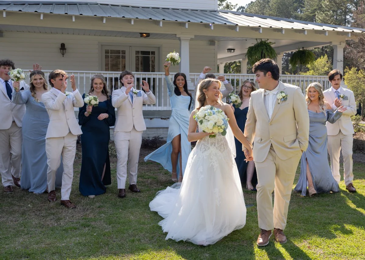 A wedding celebration with a bride and groom walking hand in hand outdoors, surrounded by their wedding party cheering and holding bouquets, in front of a white house with greenery.