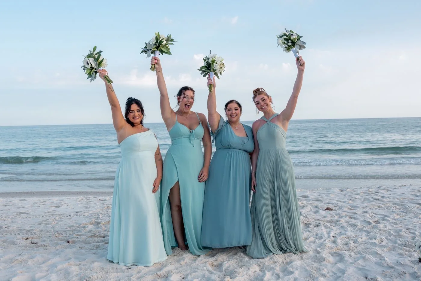 Four women in light blue dresses celebrating on a beach, holding bouquets, with the ocean and sky in the background.
