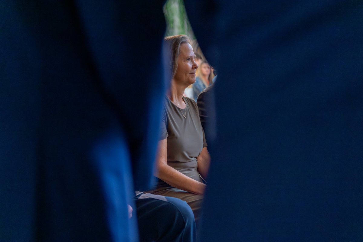 A woman sitting outdoors, framed by dark clothing or objects in the foreground, with other people blurred in the background.
