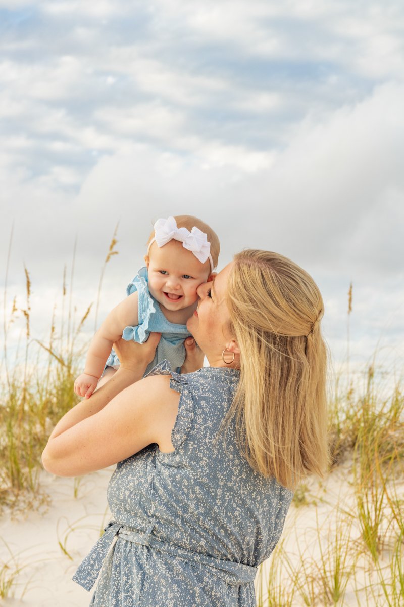 A woman holding a smiling baby girl with a white bow on a beach with sand and grass, cloudy sky in the background.