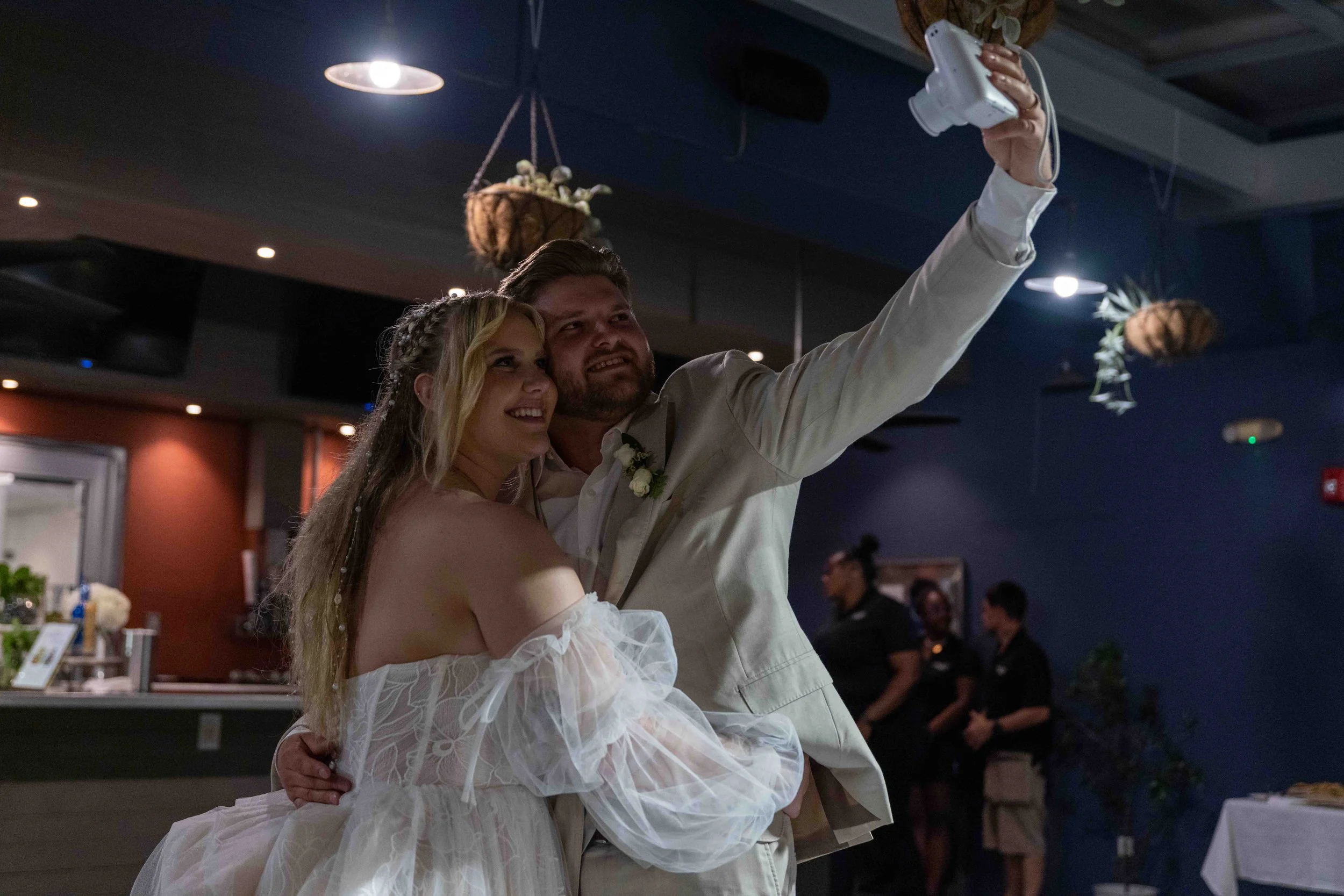 A happy bride and groom taking a selfie at their wedding reception, with guests and decorations visible in the dimly lit background.