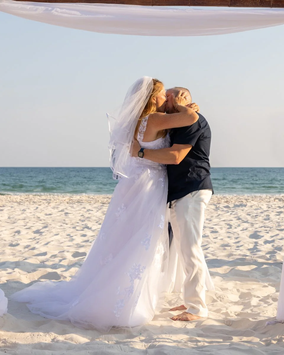 A couple kissing on the beach during their wedding ceremony, with a bride in a white dress and veil and a groom in black shirt and white pants, with ocean and clear sky in the background.